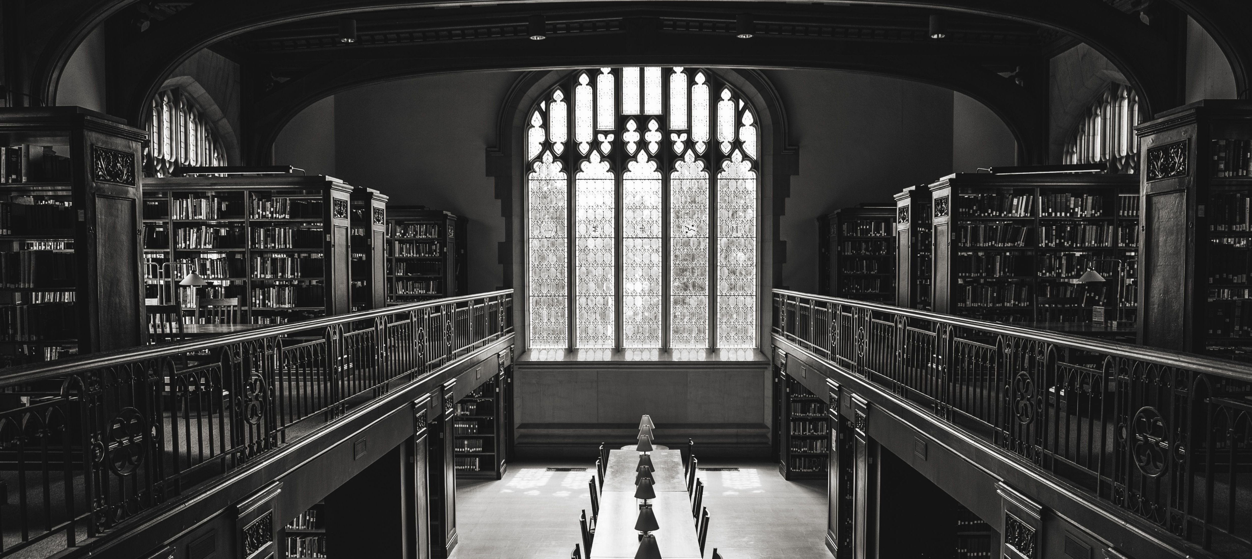 Black-and-white library interior with bookshelves, a long study table, and a large arched window